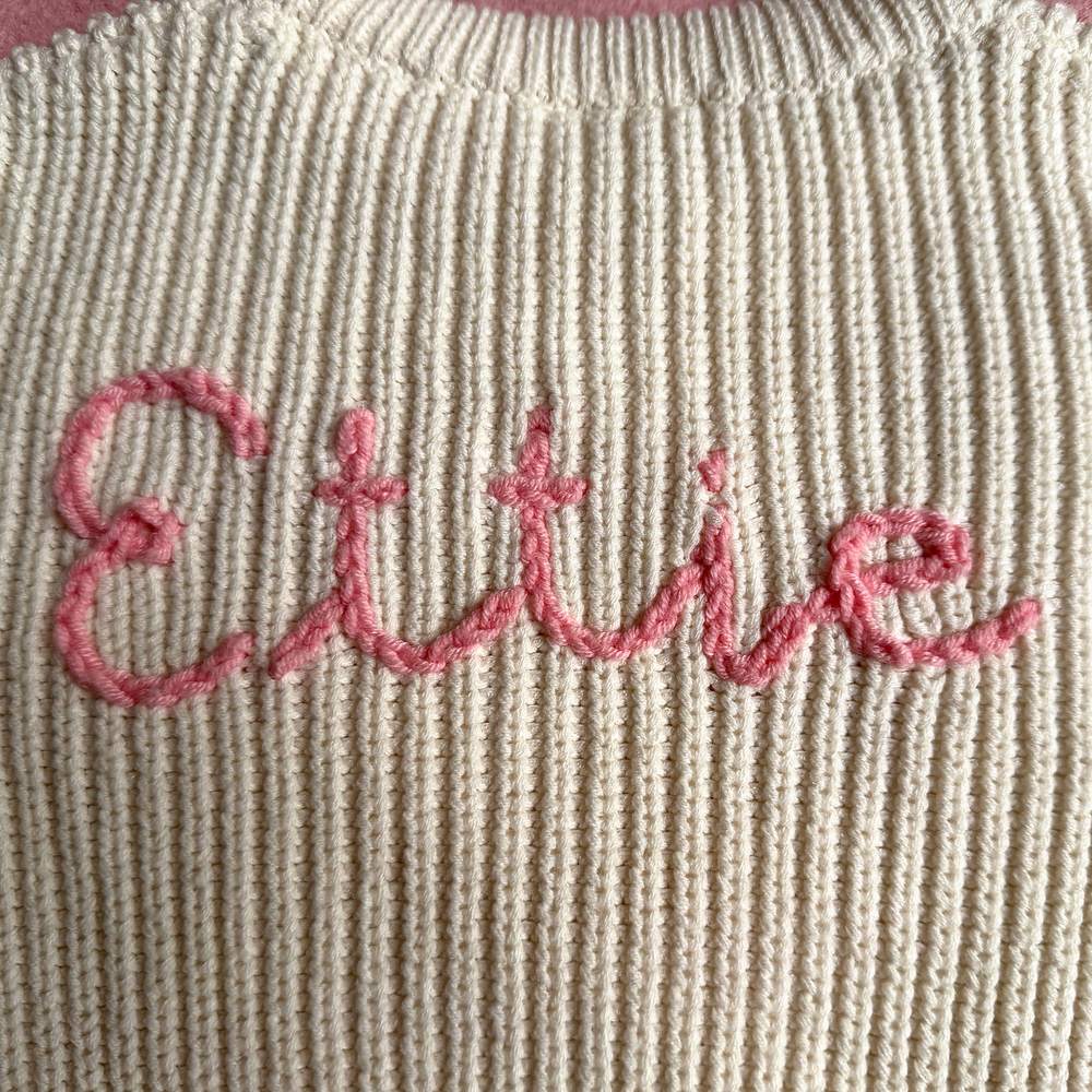 Close-up of the name Ettie hand-stitched in pink chain stitch on a cream chunky rib knit