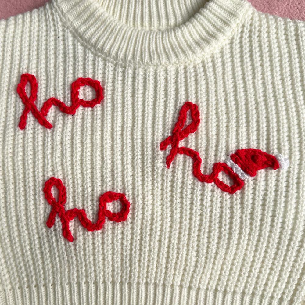 Close-up of ho ho ho hand-stitched in red chain stitch on a cream chunky knit, with a tiny red and white Santa hat on the last o
