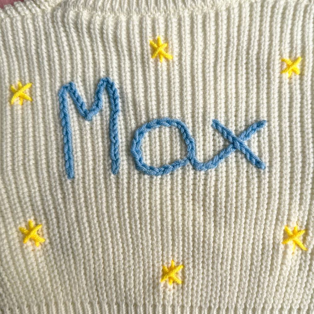Close-up of the name Max hand-stitched in blue chain stitch with yellow cross-stitch stars on a cream chunky knit